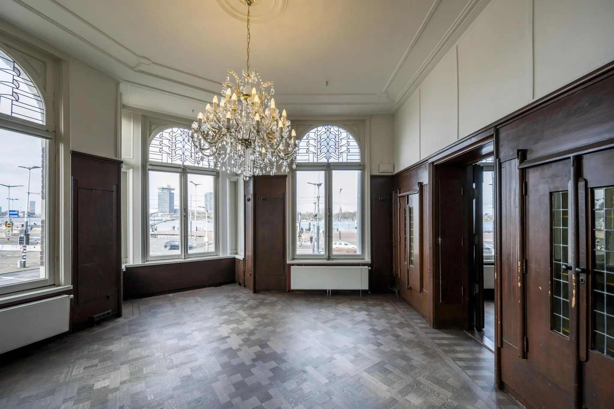 Lichte kamer aan de De Ruijterkade met houten lambrisering, grote ramen, een parketvloer en een sierlijke kroonluchter.